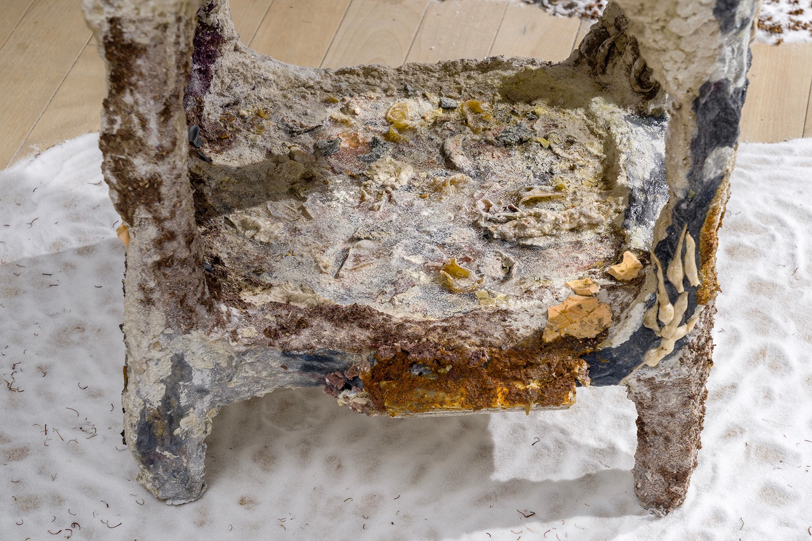 A close-up detail of a textured sculpture resembling a step or stool sits on white sand. Layered materials result in a textured, or corroded surface in shades of white, brown, gray, and yellow.