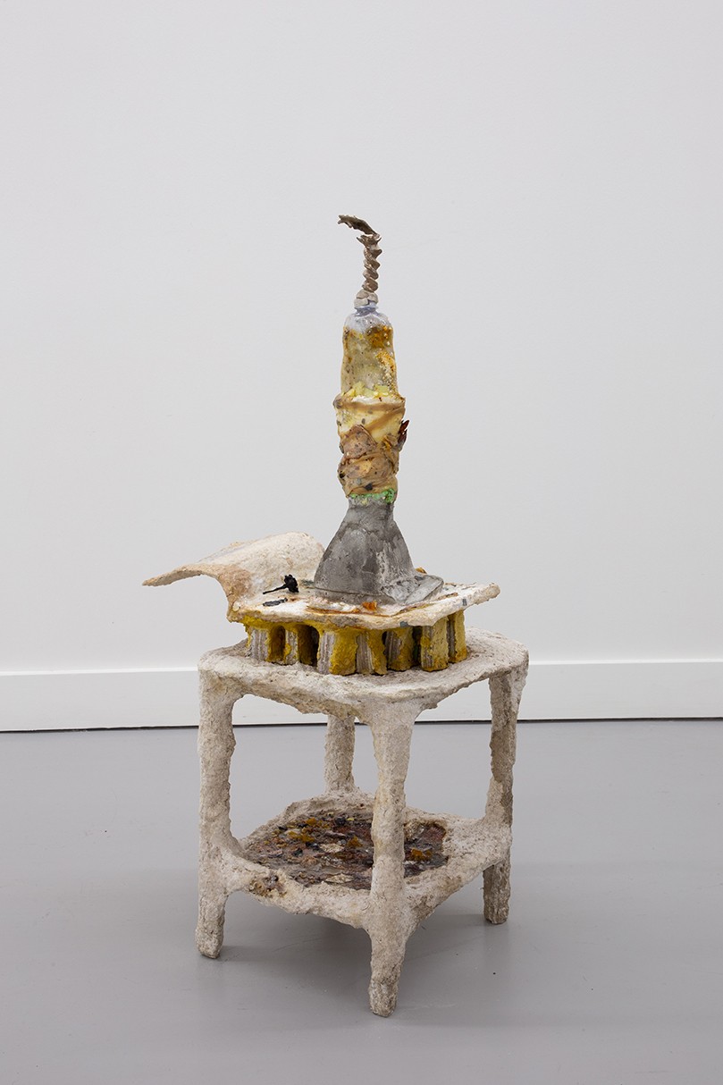 A small, sculptural object sits on a low, textured stand against a light gray background. The sculpture features a tall, slender tower made of various materials and colors, including yellow, gray, and brown. The top piece of the tower resembles a twisted corkscrew that is bent.