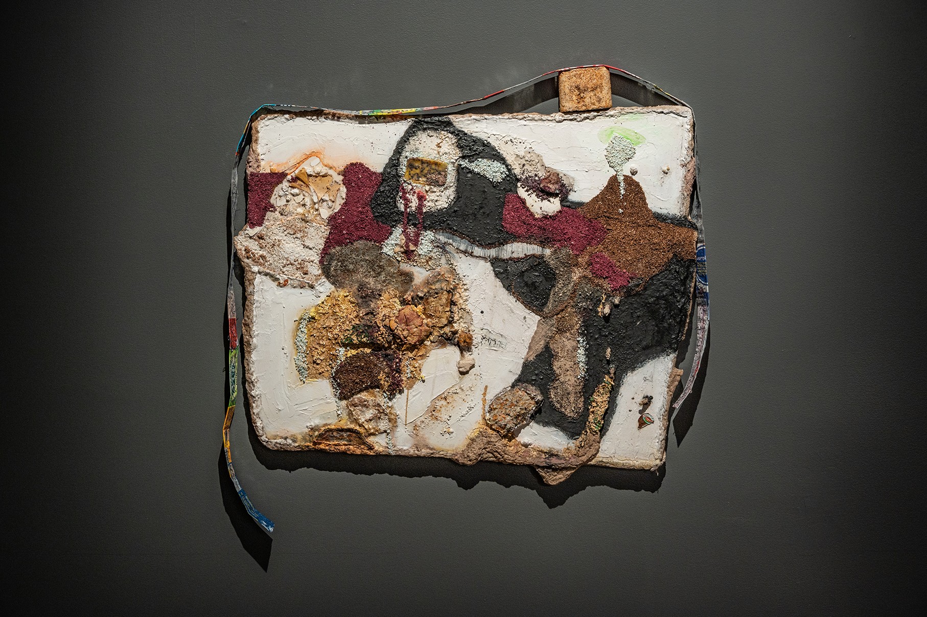 A square artwork with a textured surface is displayed against a dark gray background. The artwork features abstract figures in various colors and textures, creating a dynamic and layered composition. A thin, colorful ribbon hangs from the top left corner, and a small, rectangular object is attached to the top right. 