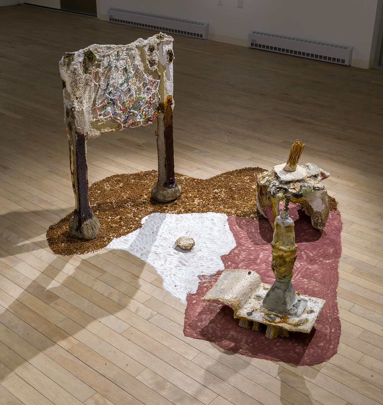 The base of the sculpture is an irregular puddle shape in three distinct textured sections in brown, white, and red. Three sculptural elements complete the composition: a tall, textured structure resembling a sign with colorful details, a smaller, table-like structure, and irregular tower, or obelisk. 