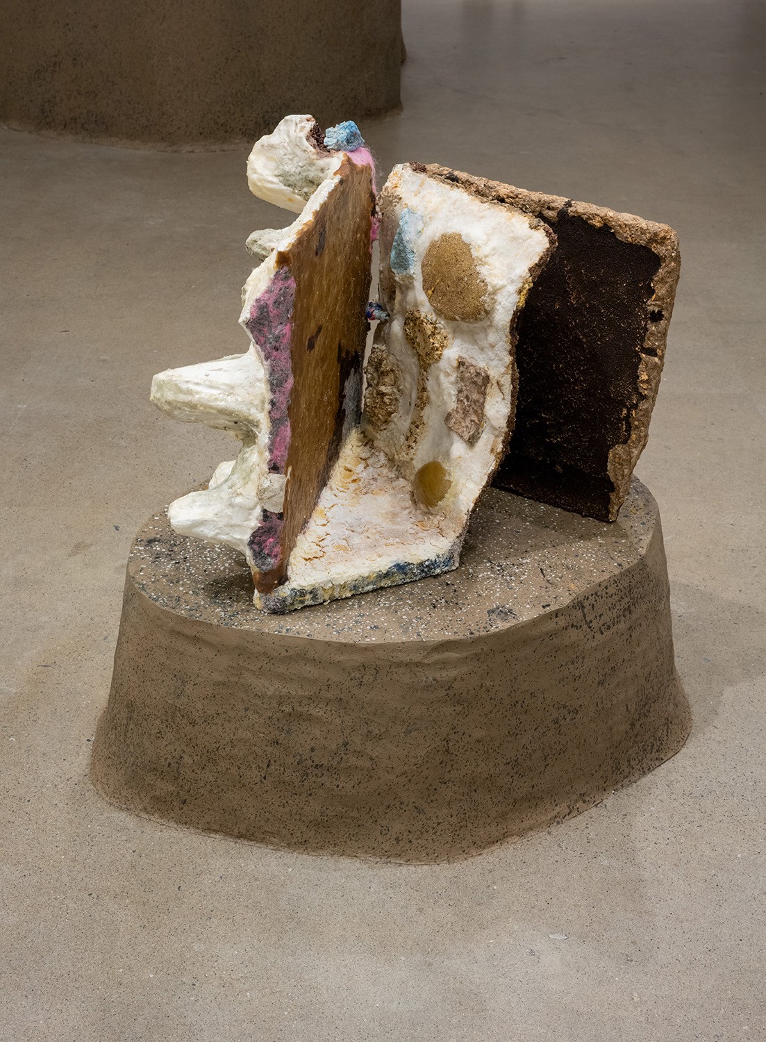A sculptural object sits on a low, earthen-colored pedestal against a gray background. The sculpture features two fragmented, book-like forms with textured surfaces and various colors, including white, brown, pink, and blue. The forms lean against each other, casting shadows on itself.