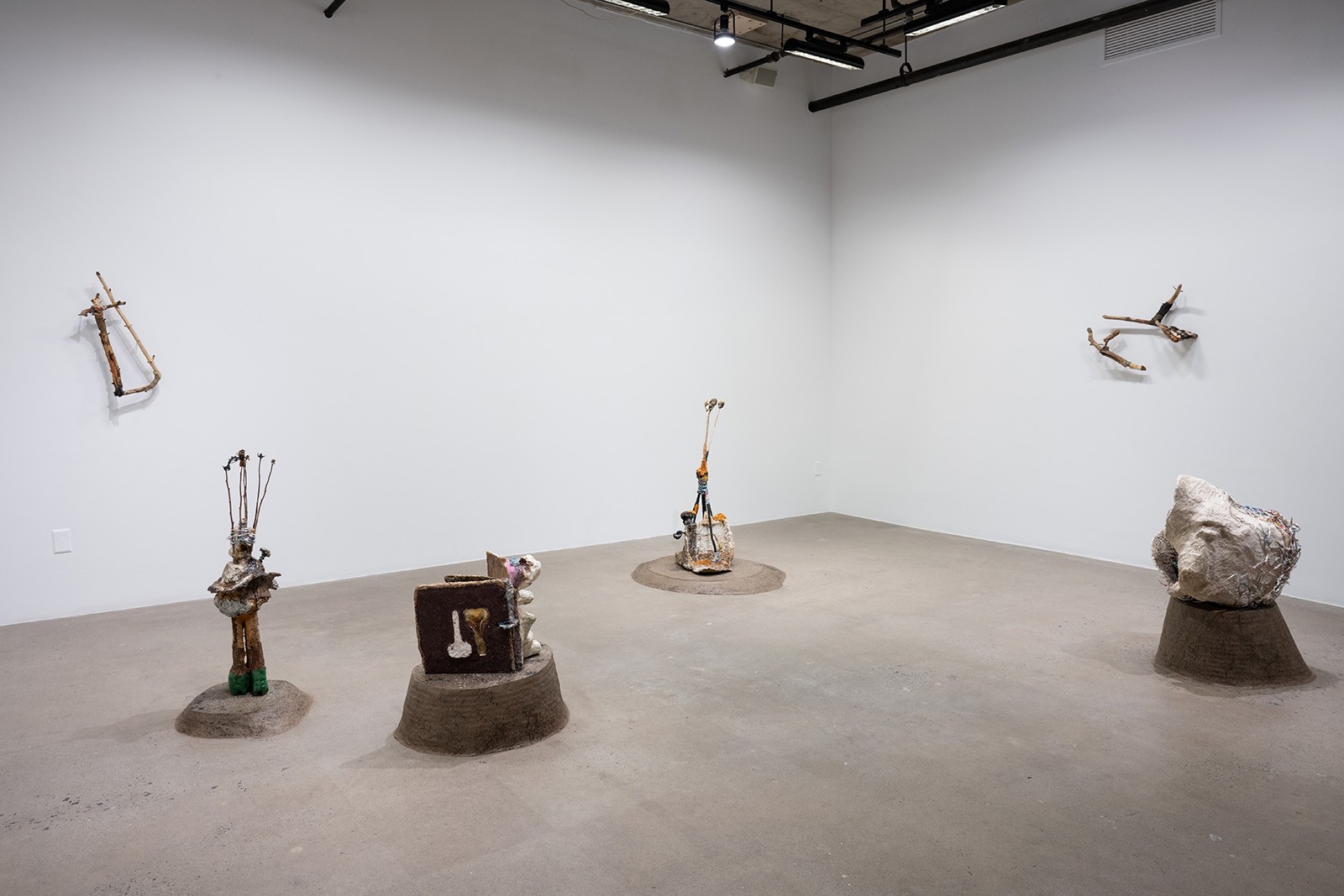 A wide shot of a gallery space with white walls and a light gray floor features several sculptural objects. The sculptures vary in size and material, including wood, metal, and ceramic. They are placed at different points around the room, creating a sense of space and distance. 