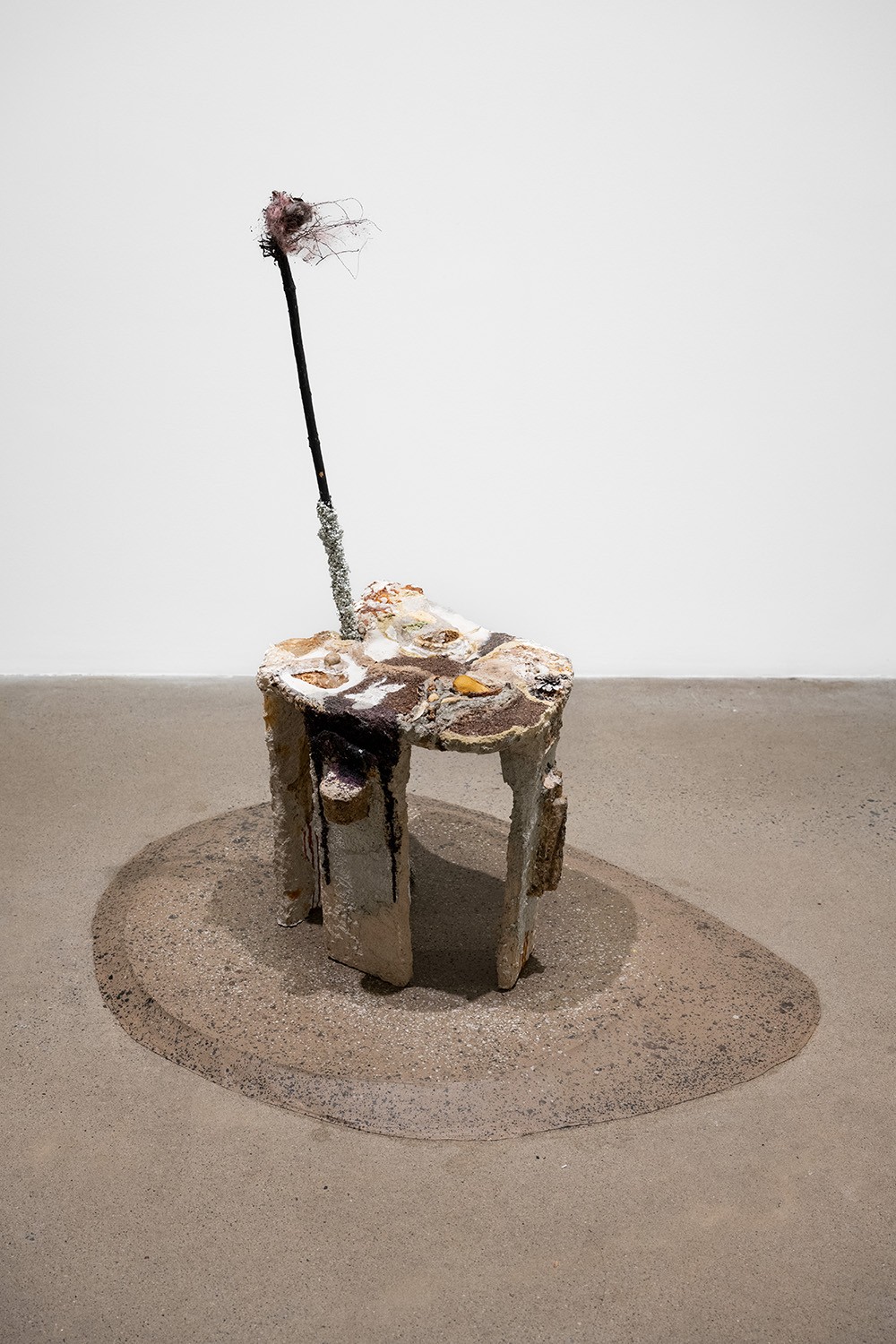 A small, whimsical sculpture stands on a circular, textured base against a light gray background. The sculpture features a rough, table-like structure with a tall, slender stick protruding from it. The stick is adorned with a small, pink object and thin, wire-like elements. 