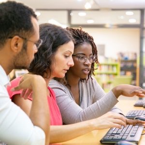 Two women and one man sit at a table looking at a computer screen. 