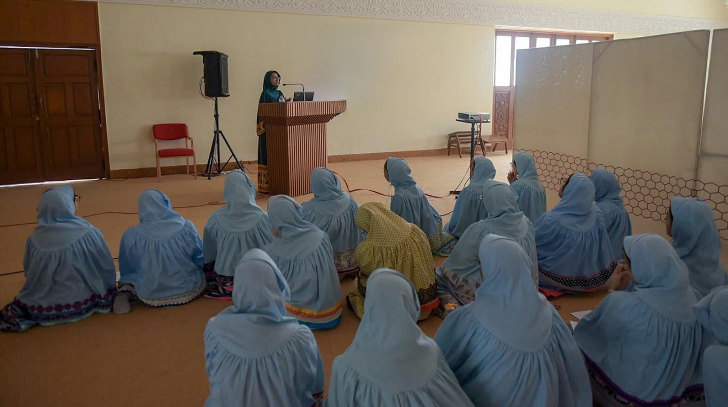 A woman wearing a green hijab and traditional Pakistani clothing stands at a wooden podium in front of a group of eighteen women seated on the ground of a large room with beige walls and brown floor. The women on the floor are all wearing blue hijabs except for one woman in the center who is wearing yellow.
