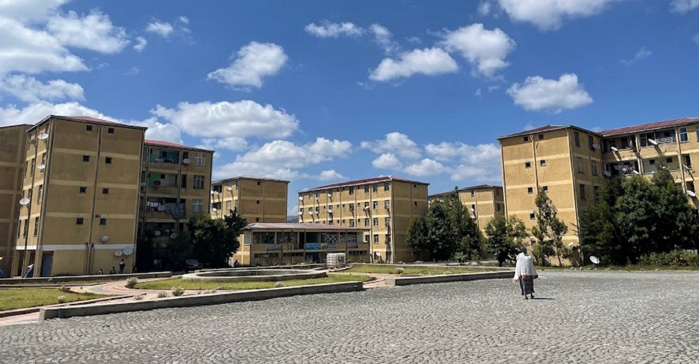 A few multi-storied apartment buildings with sand-couloured walls dominate middle ground. Above the buildings, a blue sky with scattered fluffy white clouds. In the foreground, a woman wearing a white shawl walks across a cobblestone square towards the buildings.