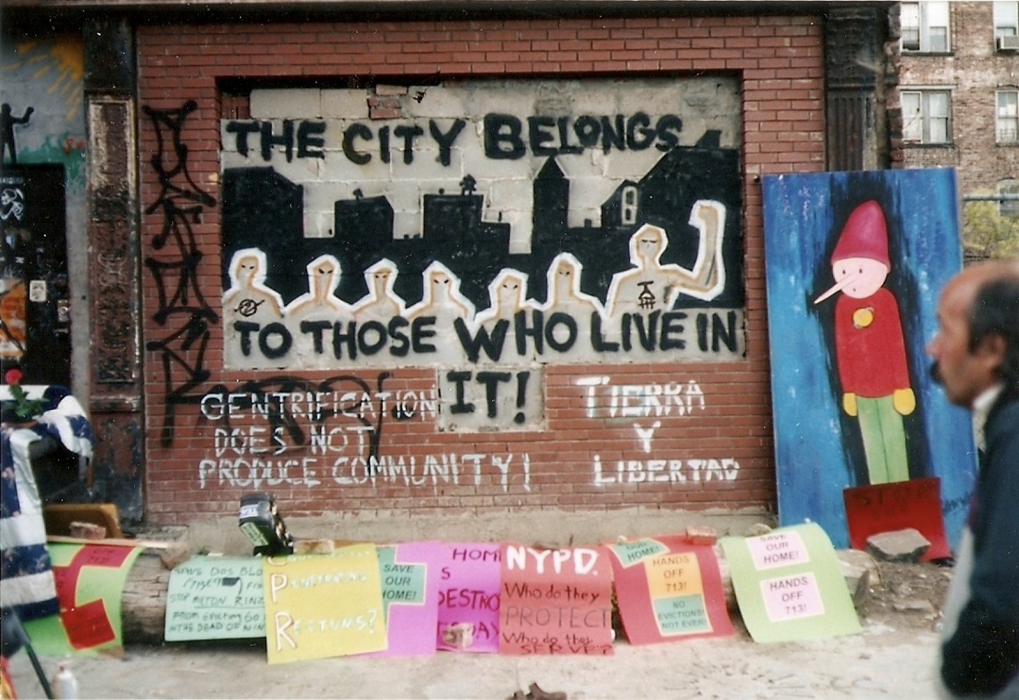 A red-bricked wall with a rectangular cut out at the center with grey bricks. The following words are painted in black capital letters “THE CITY BELONGS TO THOSE WHO LIVE IN IT!”. Below these words on the red brick, the following messages are painted in white letters: “Gentrification does not produce community!” and “Tierra Y Libertad”. A series of handmade posters line the ground in front of the mural, while a vertical painting depicting what appears to be Pinocchio is propped against the wall to the right.