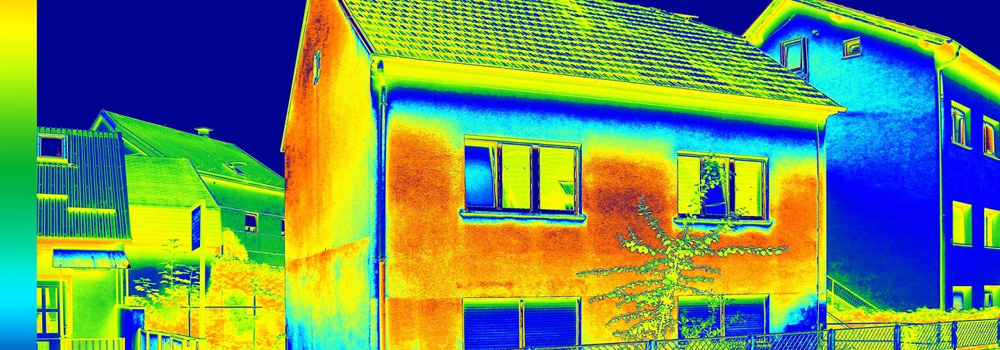 A thermal image of a two-storied residential house with other houses to the side and in the background. The featured house is shown to have the most warmth centered on the front and left side walls, shown in yellow and orange. The cooler areas are along the edge of the roof and the lowest visible section of the house and are shown in blue.