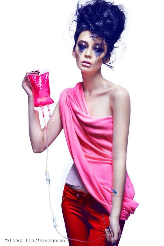 A woman in a pink top with dark eye makeup streaked across her face holds up a blood bag connected to her forearm, containing vivid pink liquid.