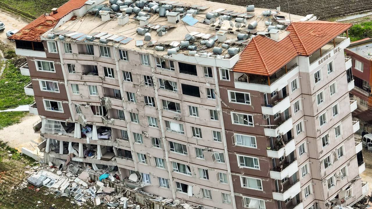 A high-rise residential building leaning dramatically with its ground floor destroyed after an earthquake, showing major structural damage.