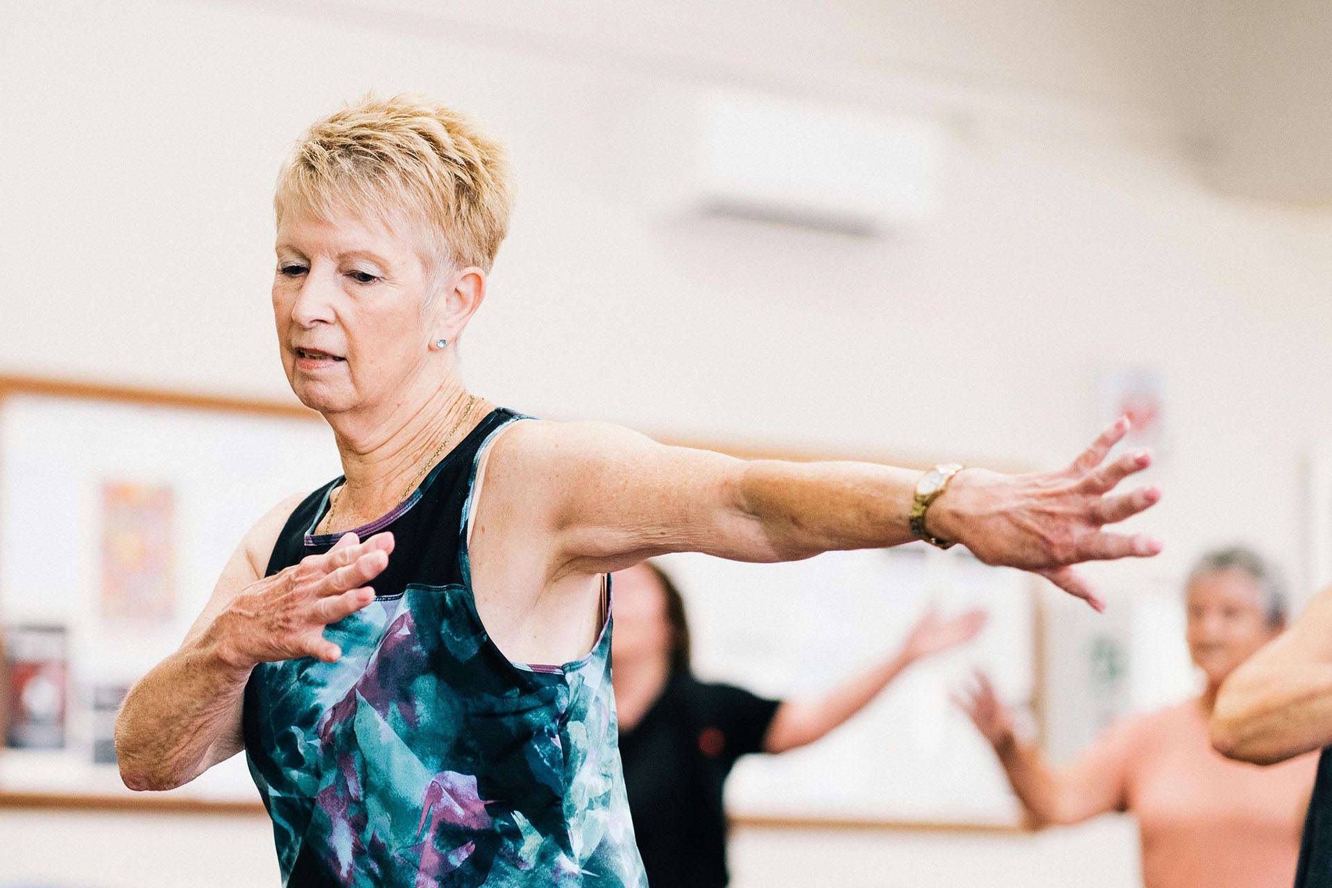 An older woman with short blonde hair and a blue/purple patterned top reaches to the side during a dance class.