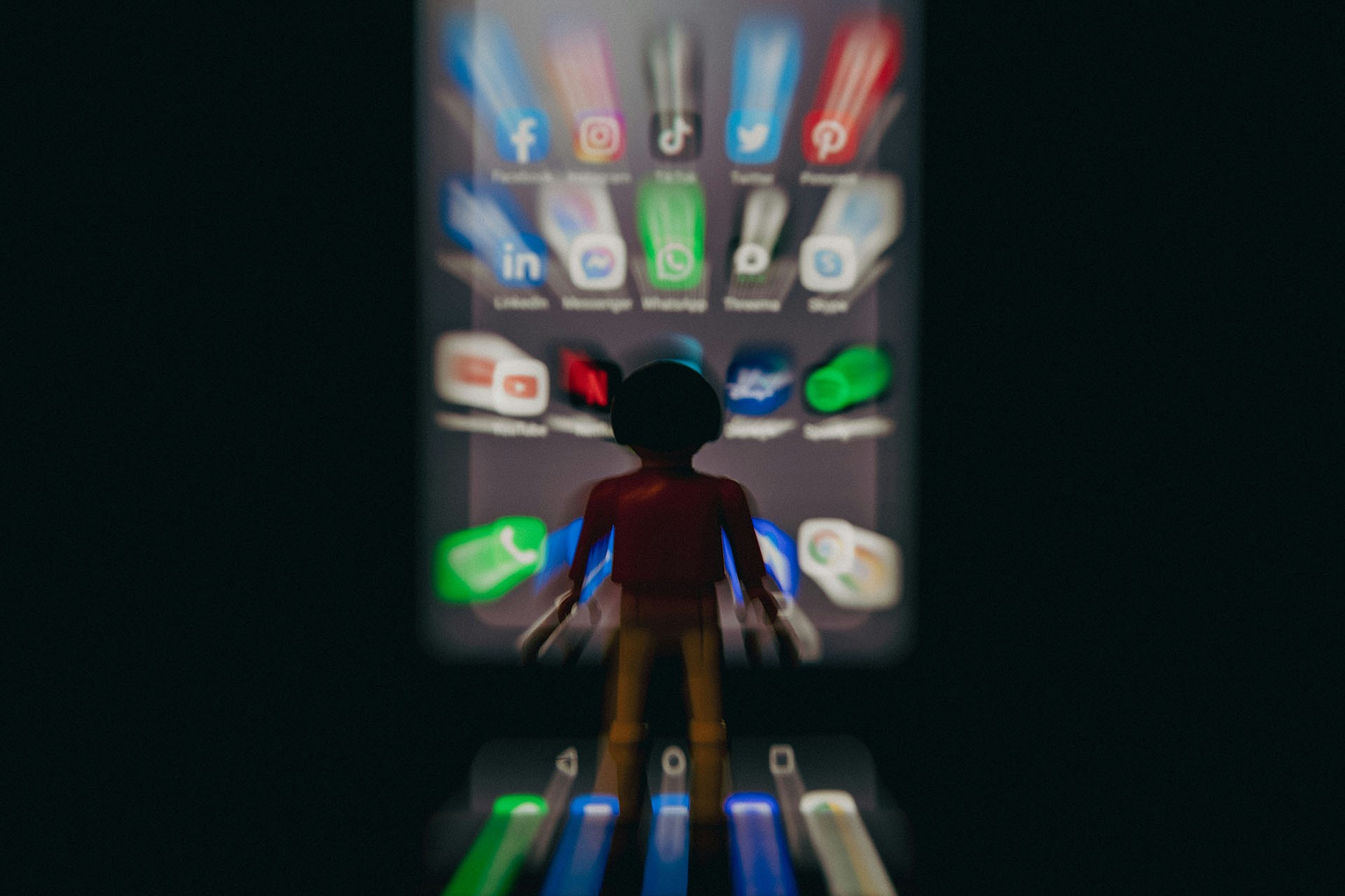 A toy figure stands in front of a large smartphone screen displaying various social media app icons, including Facebook, Instagram, TikTok, and Twitter. The icons appear blurred with motion effects, creating a visual impression of being overwhelmed or absorbed by digital content.