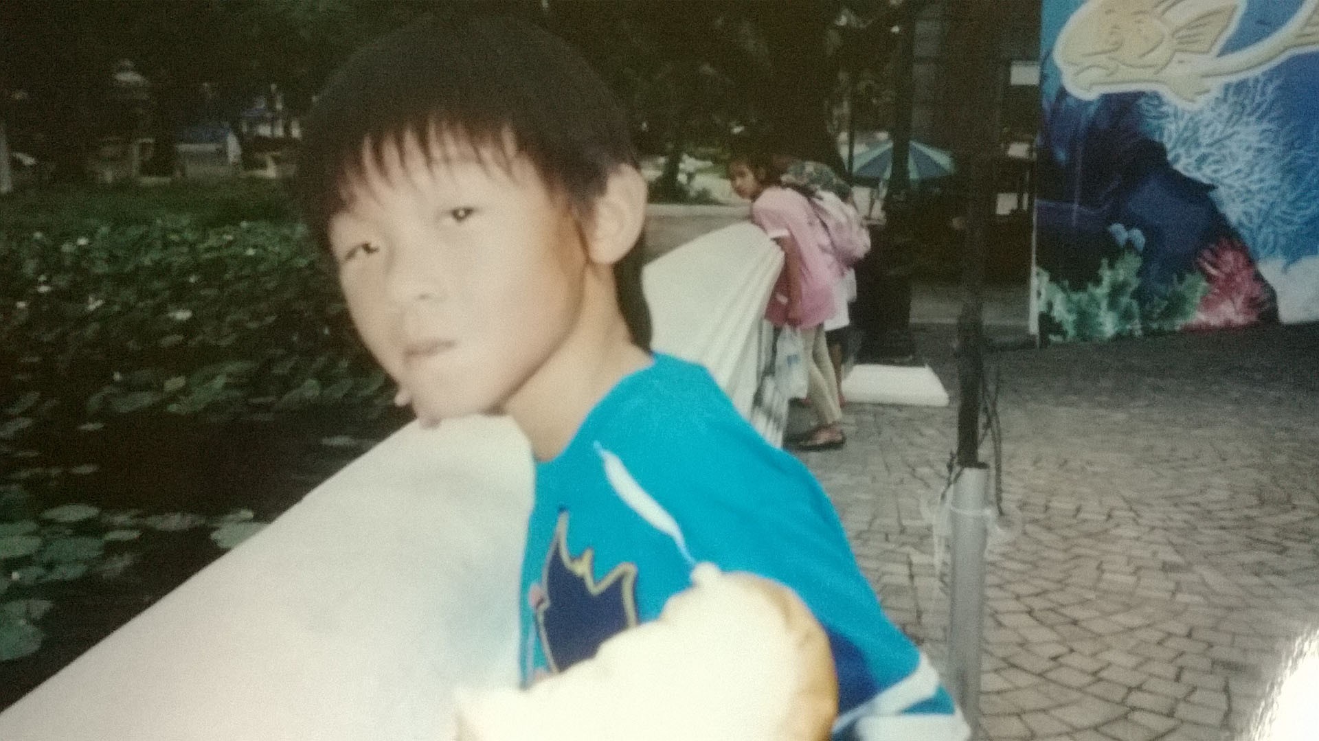A young boy in a blue shirt peers over a concrete fence that overlooks a pond with lily pads. He stares into the direction of the camera but not directly, looking beyond.