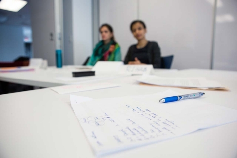 A pen on a piece of paper with scribbled notes on it. A pair of students look on attentively in the background.