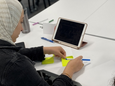 Student seated at a table with a tablet in front of her, adding post-it notes to a blank page.