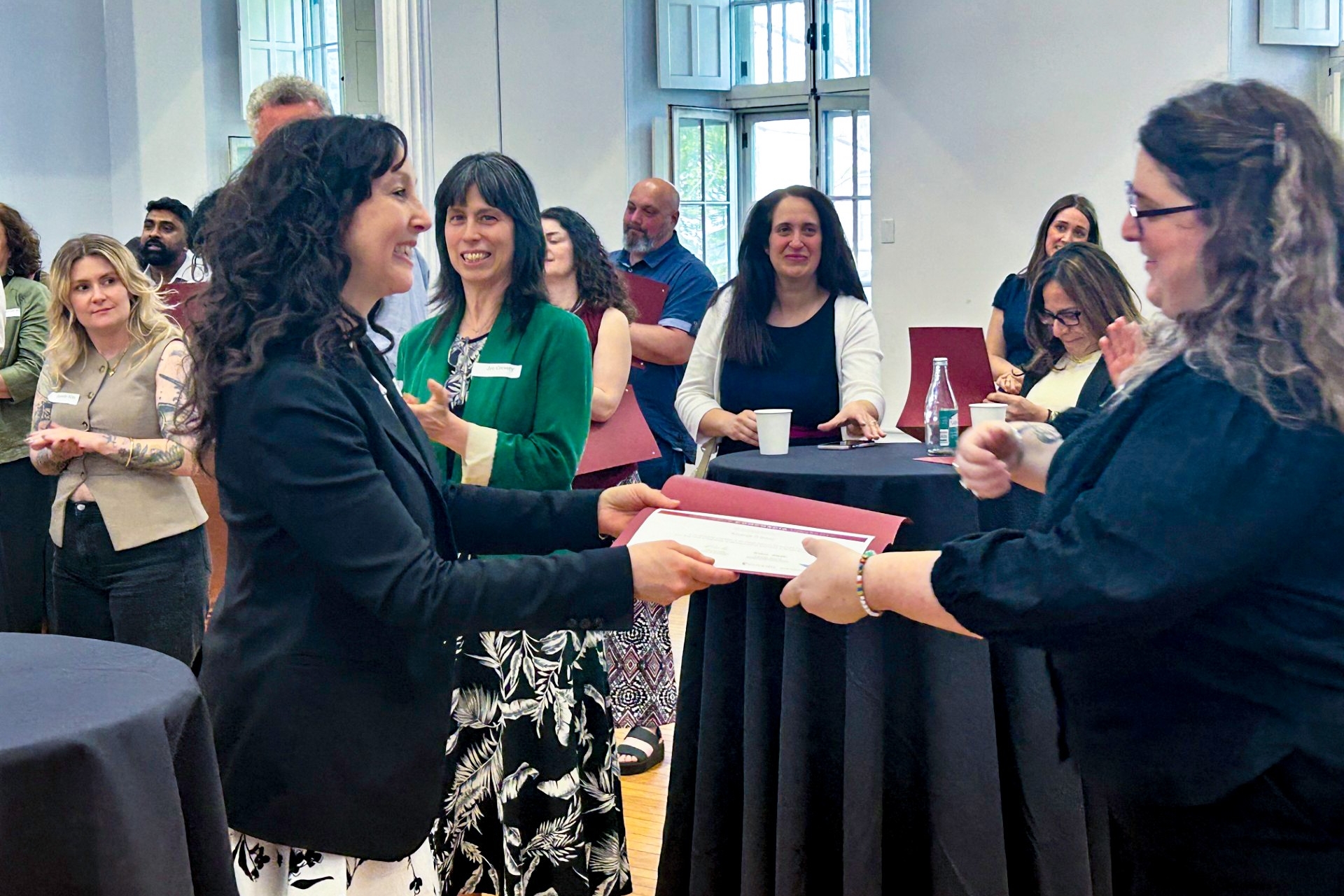 A participant of the CRDP program receives her diploma from the program's facilitator at the celebration cocktail event, in a room full with other participants clapping.