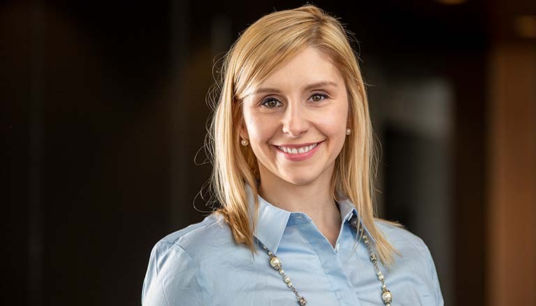 Young, smiling woman with long, blonde hair, a blue shirt and a silver and pearl necklace.