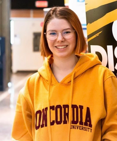 A young woman in a yellow Concordia hoodie smiles confidently.