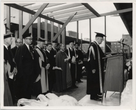 Group of people in ceremonial caps and growns stand behind a speaker at a podium