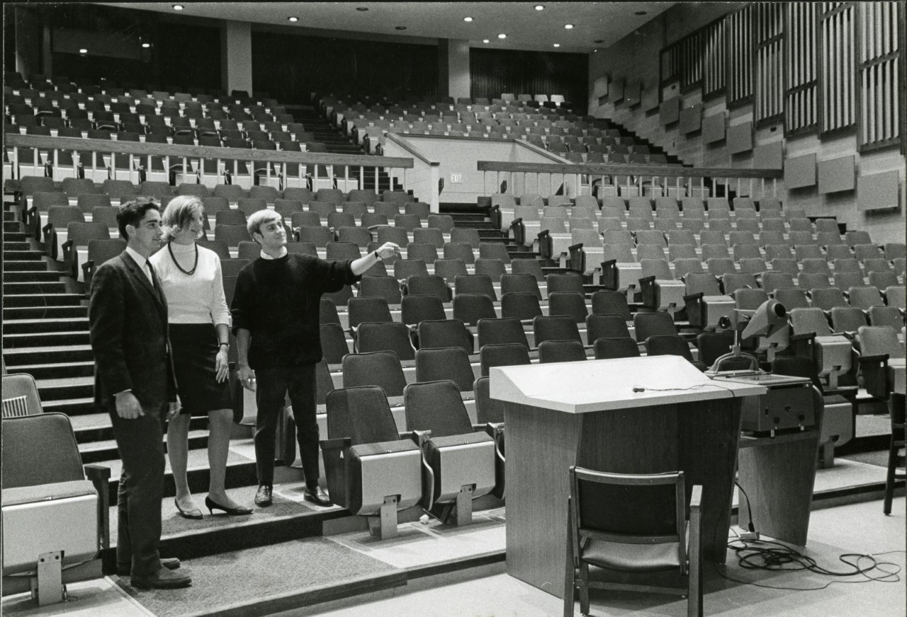 People looking toward the podium of an auditorium, one of them pointing as if explaining something