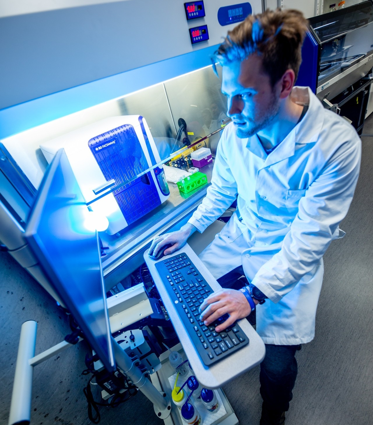 A scientist in a lab coat uses a computer keyboard while examining a blue-lit device in a laboratory setting.