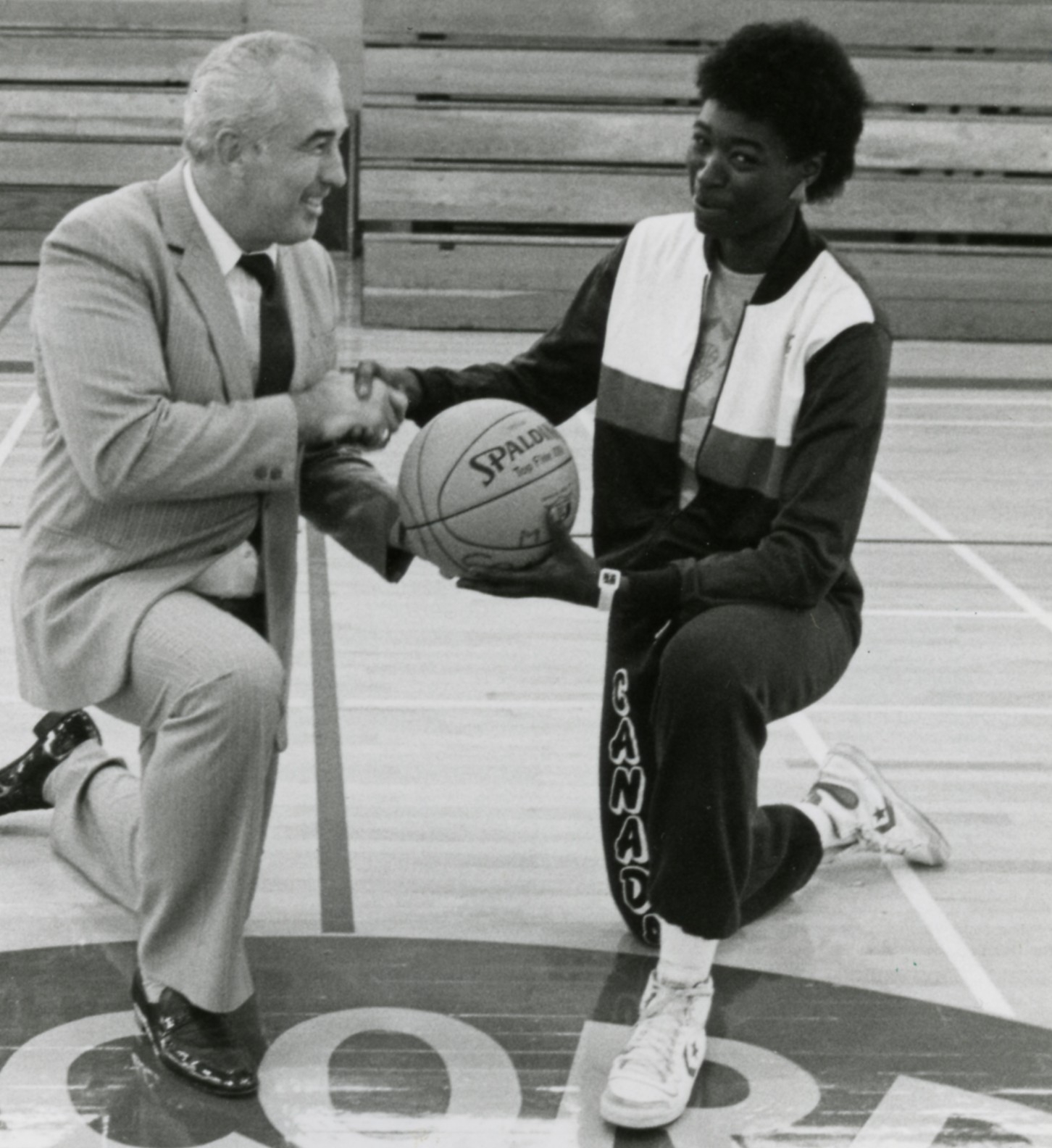 A older man and black student athlete posing for a photo. They're shaking hands and holding a basketball.