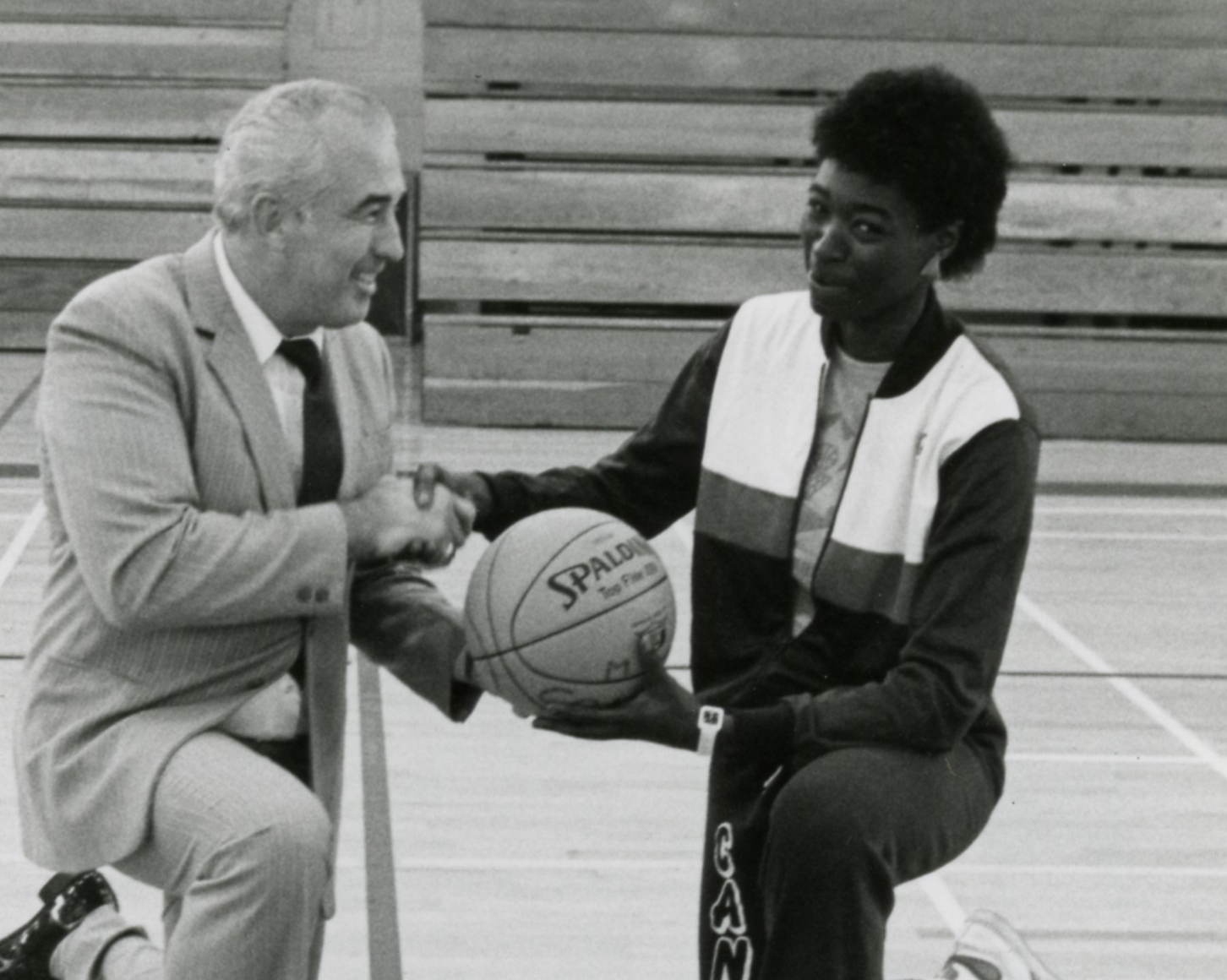 A older white man in a suit shaking the hand of a black student athlete. 