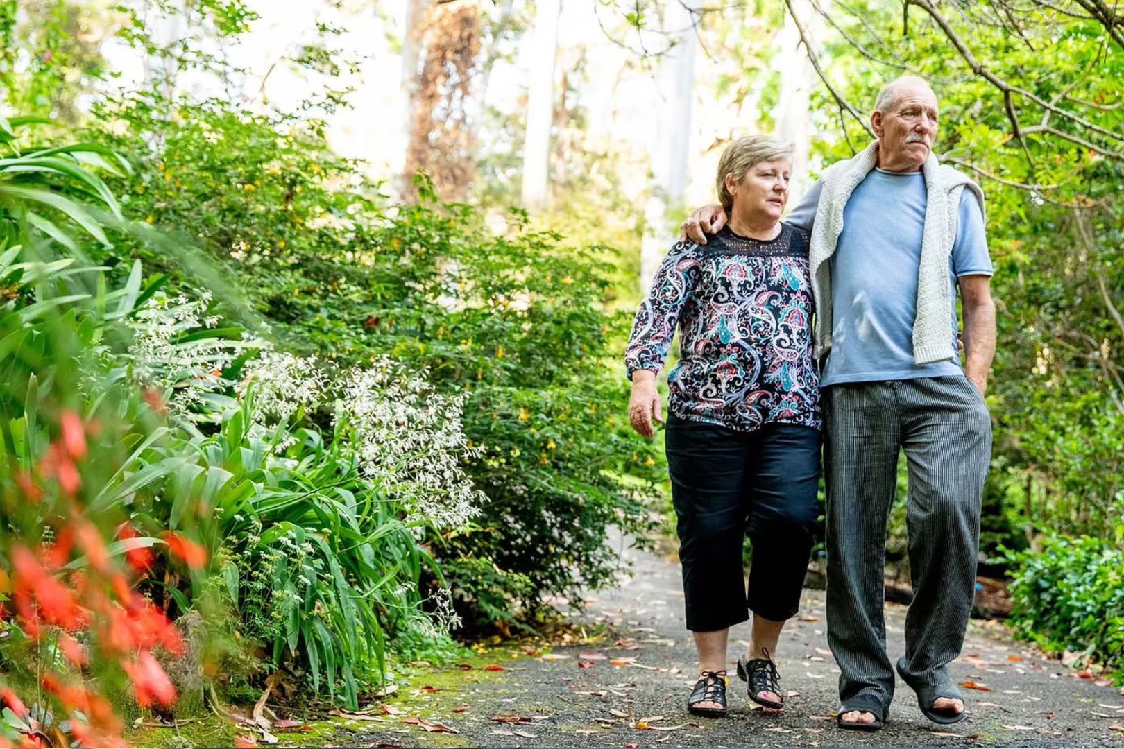 A couple of older adults, the man with his hand around the woman's shoulder, walk on a lush green nature path.