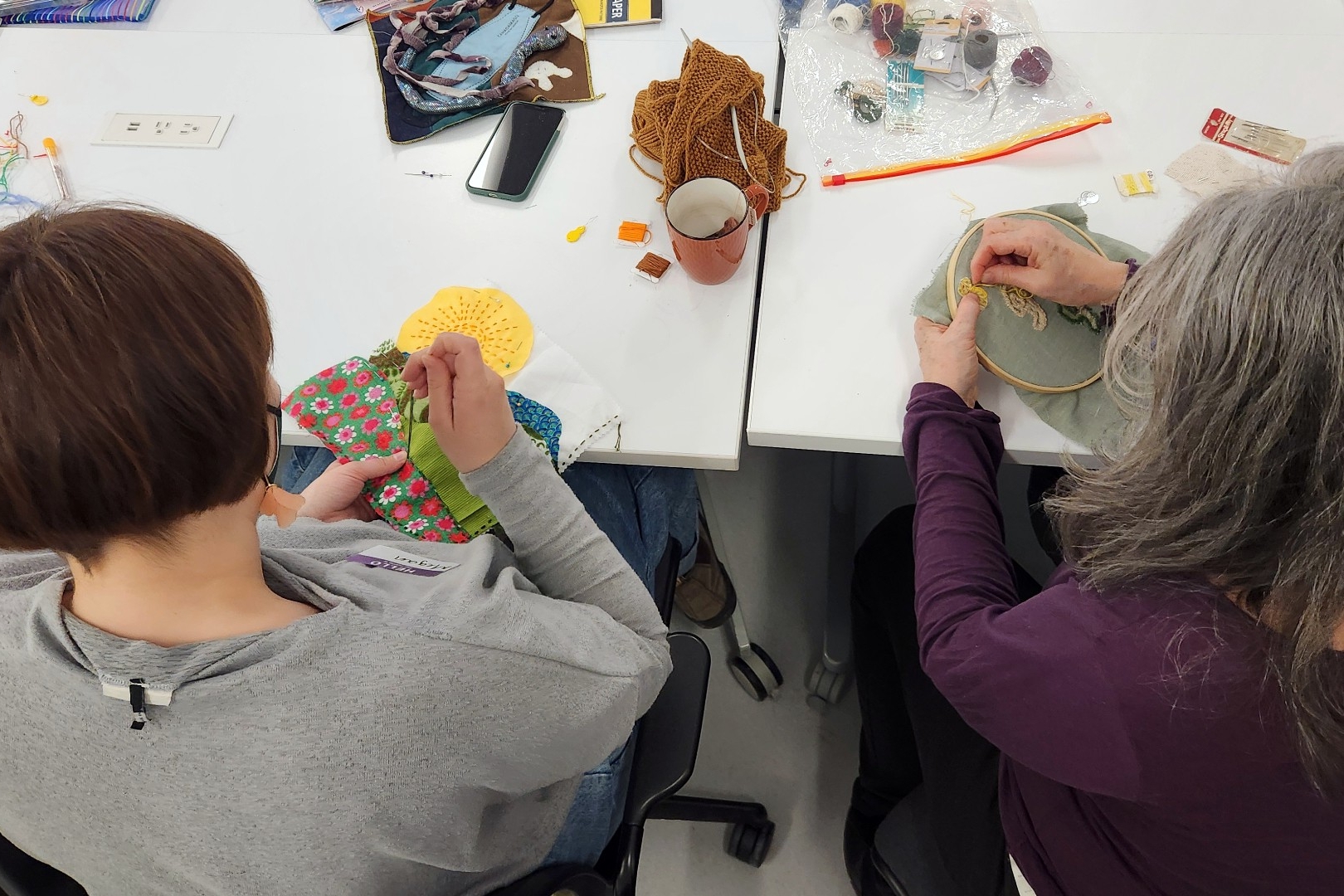 A young woman and older woman sit side by side at a table working on separate embroidery projects.