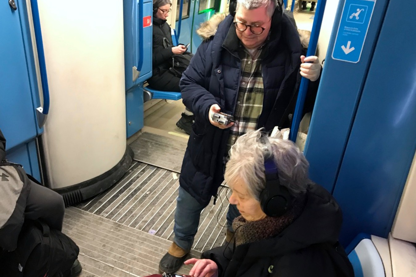 Two older adults in winter clothing, one standing and one sitting down, are on a Montreal metro train with headphones on and recording devices in their hands.