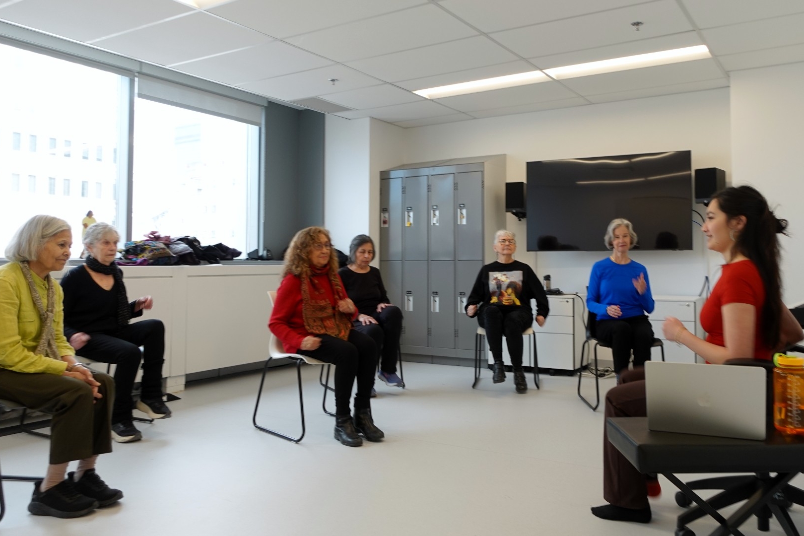 Emma Chen leads a dance exercise class. Emma is younger and sitting in a chair surrounded by 6 women sitting in chairs circling her waiting for instruction.