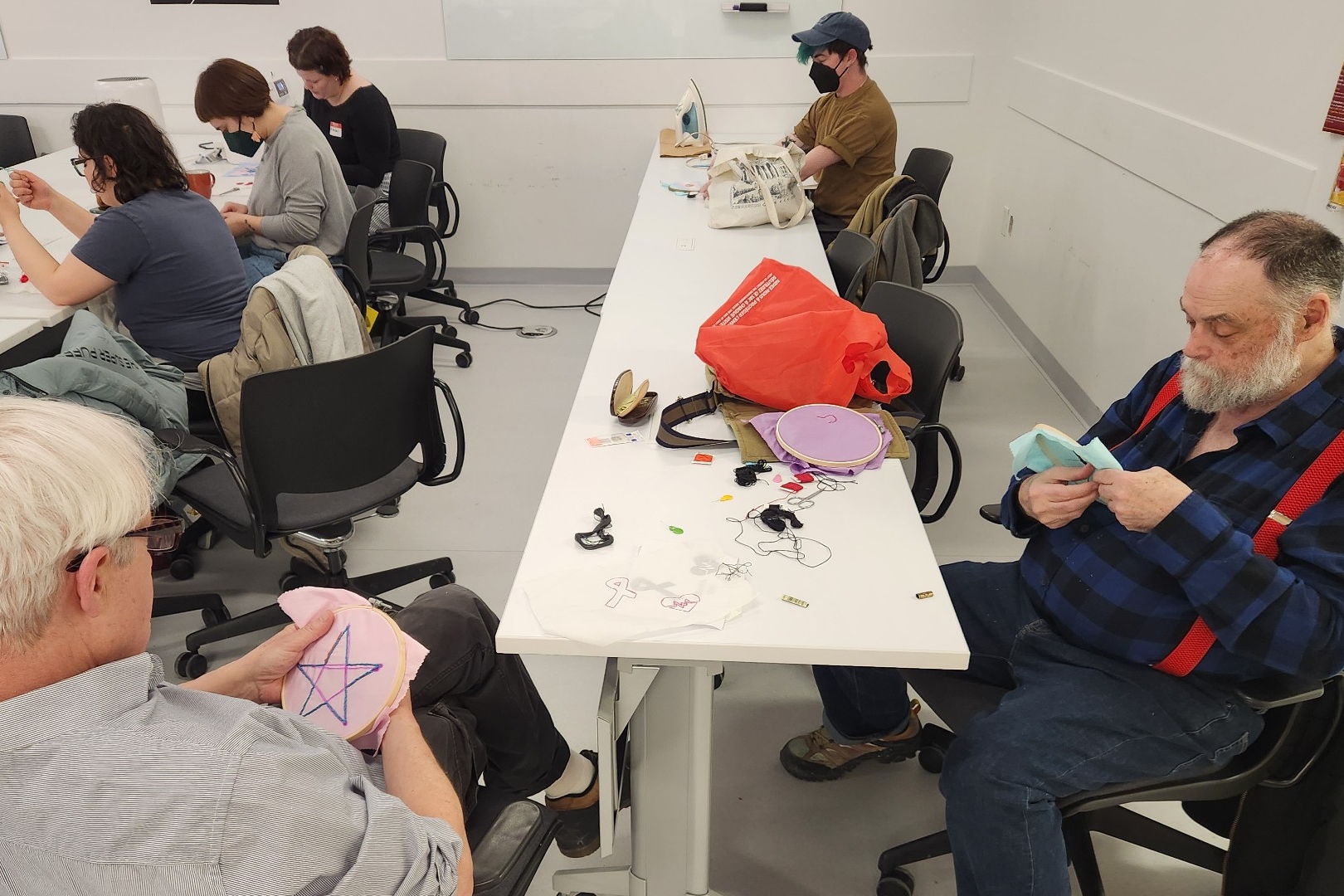A room full of people of various ages sit around tables together working on embroidery projects.