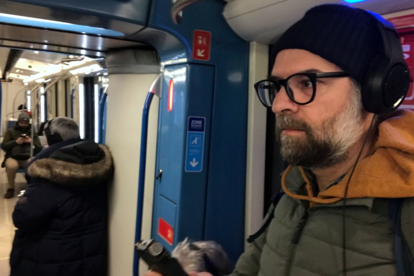 A man with a greying beard, black framed glasses, and a toque stand in the Montreal metro train with headphones on and a recording device held up in his hand.