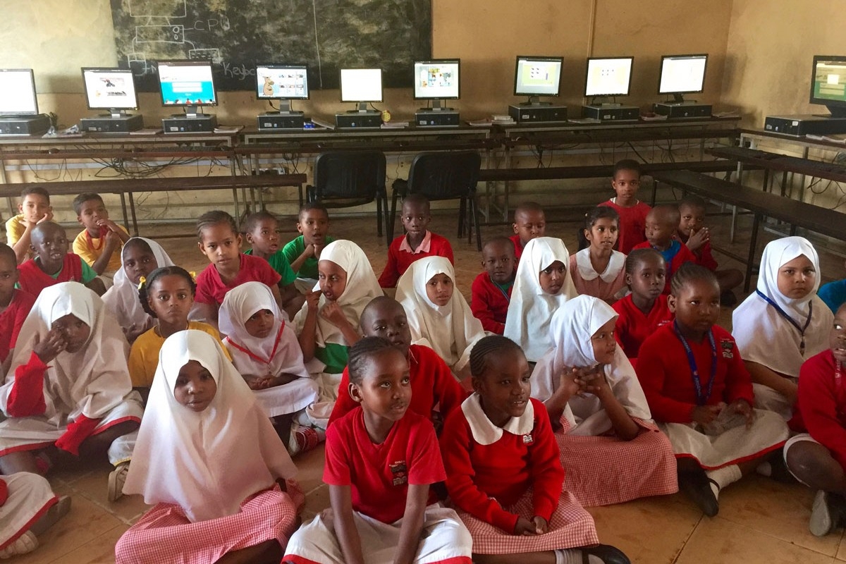 A group of children, wearing mostly white and red outfits, sit on the floor attentively. 