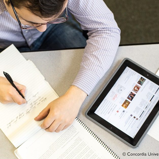 a man writes on notepaper while sitting at a desk with a notebook and tablet