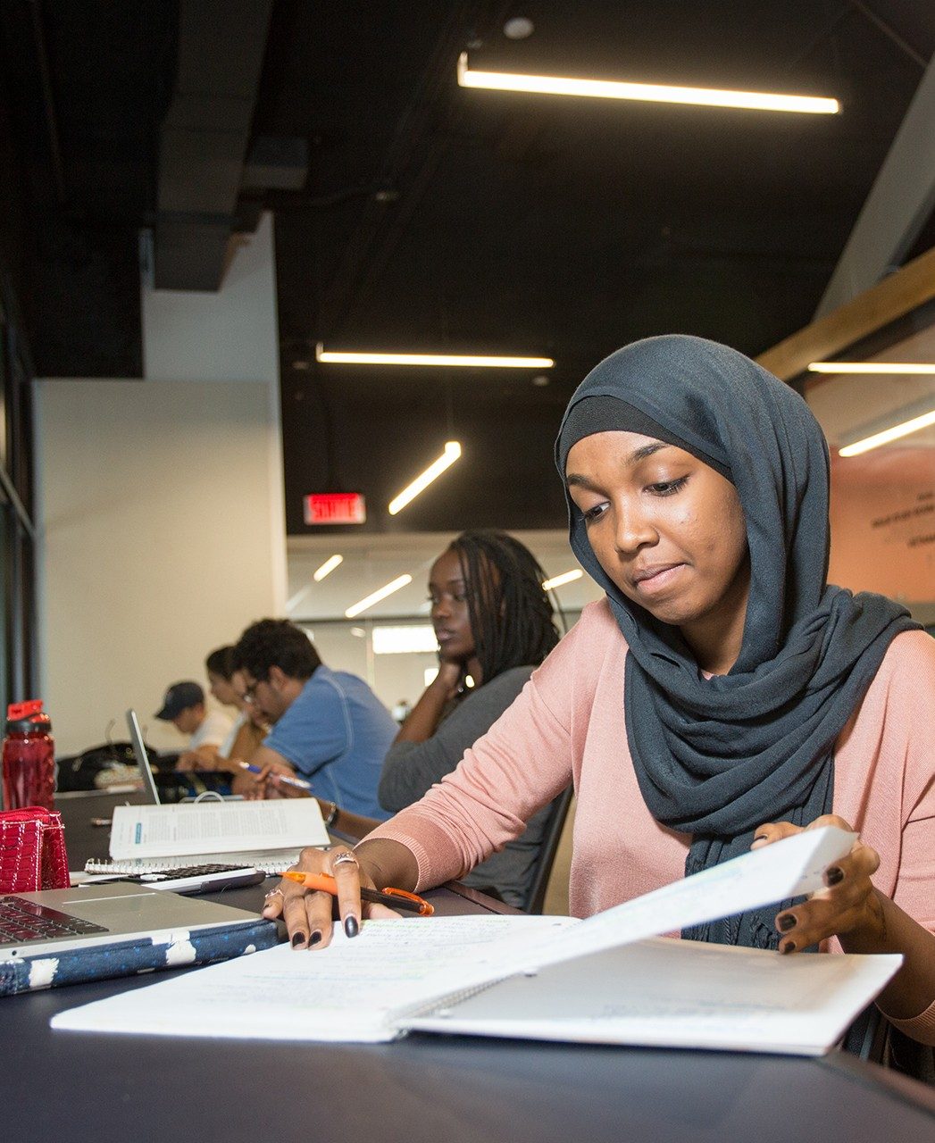 A student wearing a hijab reads a page in a notebook and other students study nearby A student wearing a hijab reads a page in a notebook and other students study nearby