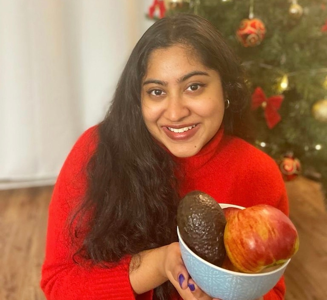 A female with long brown hair sits in front of a Christmas tree while holding a bowl of fruit.