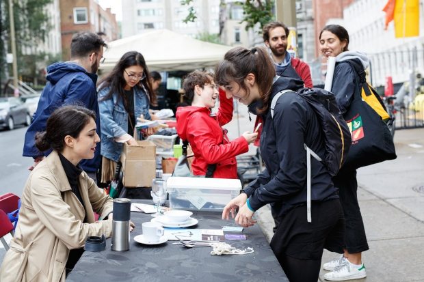 People at an outdoor event table, engaging in conversation and handling materials. There's a mix of casual attire and activity in an urban setting.