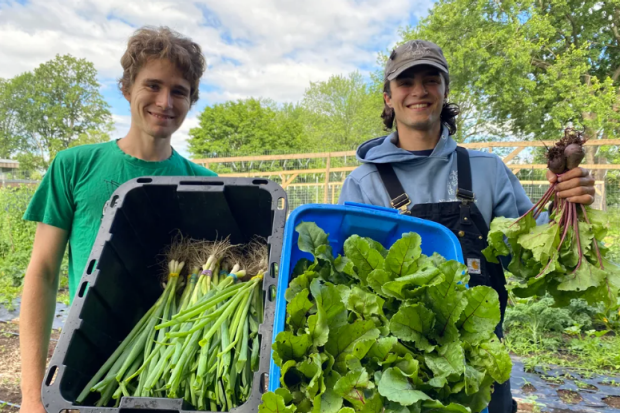 Two smiling people hold bins of fresh produce in front of a garden at Loyola