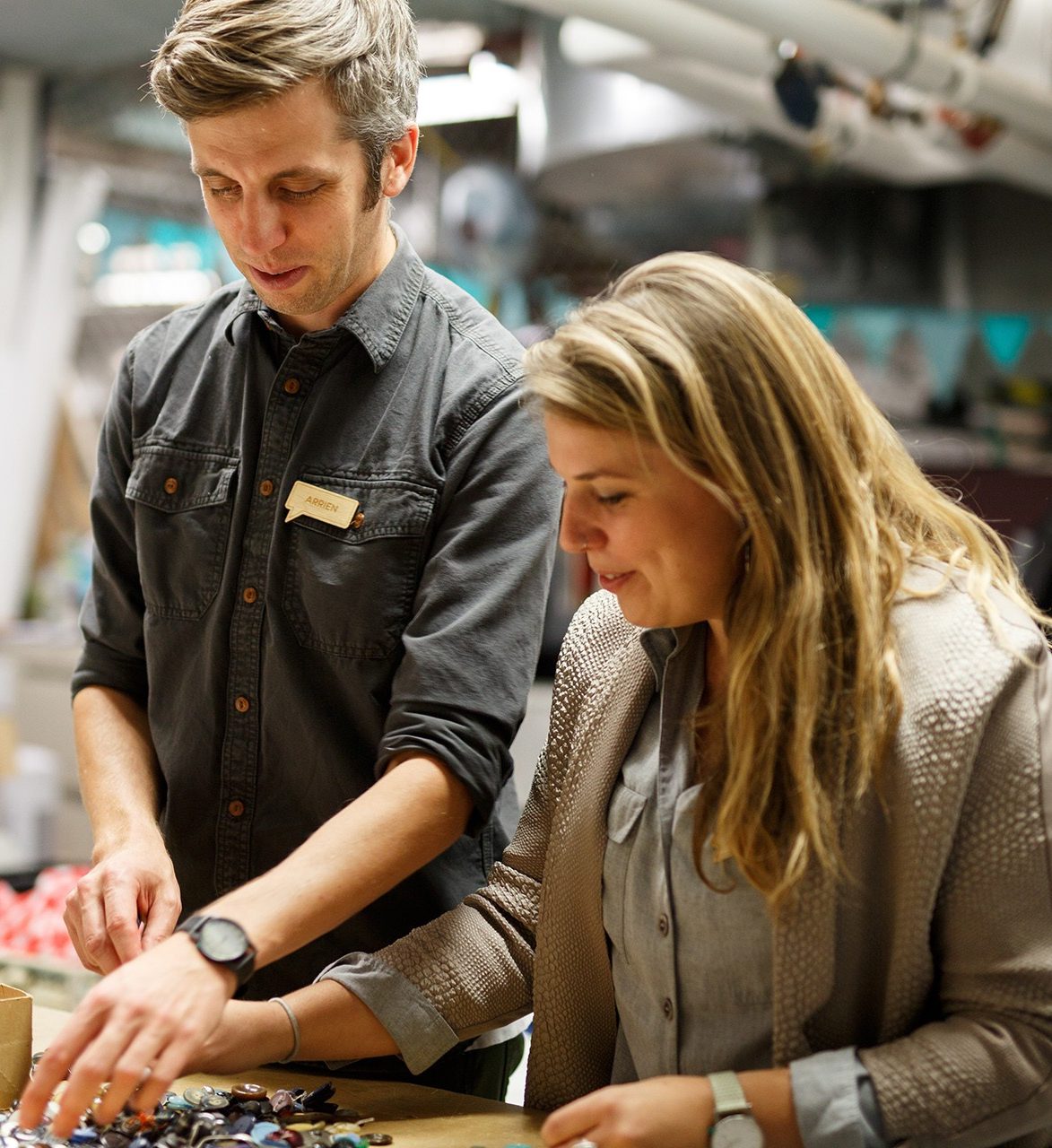 a man and a woman stand at a table sorting small objects against an industrial backdrop