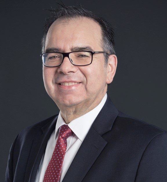 A smiling man wearing a suit and glasses smiles at the camera