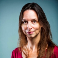 A woman with long brown hair wearing a red shirt stands in front of a plain blue background, looking at the camera with a neutral expression. 