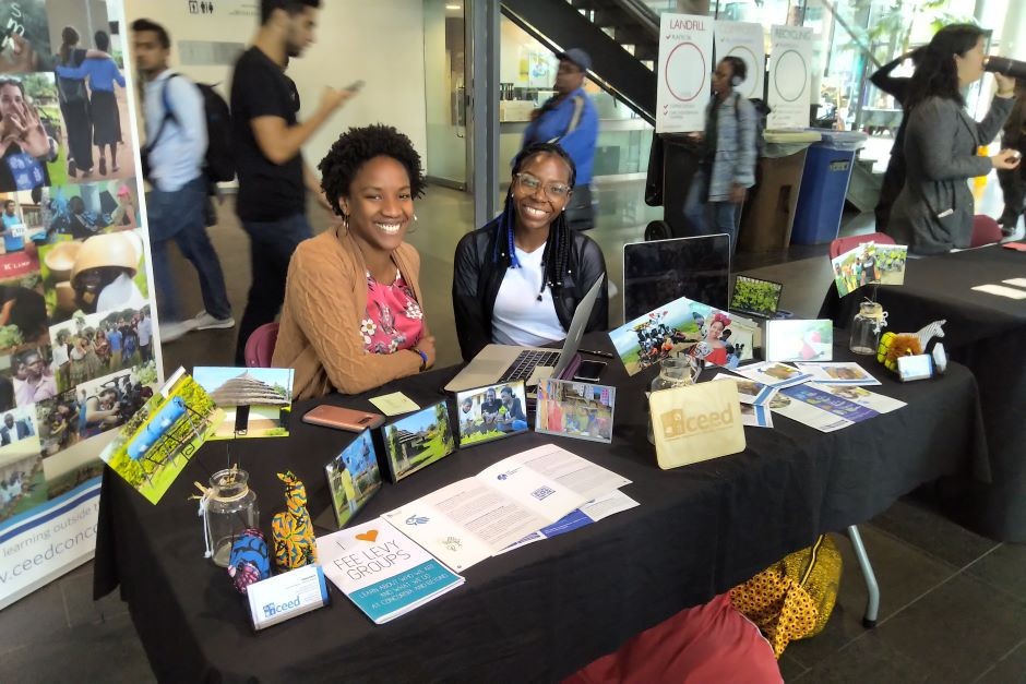 Executive Director, Hawa Keita, and Internship Coordinator, Josie Fomé, of Ceed Concordia sit at a table covered in materials depicting the activities of CEED. In the background, people walk past and a CEED banner stands.