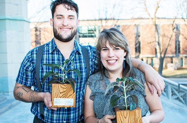 Two smiling people stand next two each other, one with his arm around the other. They are both holding Sustainability Champion Awards in the shape of planters holding seedlings.