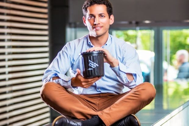 A person sits cross-legged holding a small black container indoors, wearing a light blue shirt and brown pants.