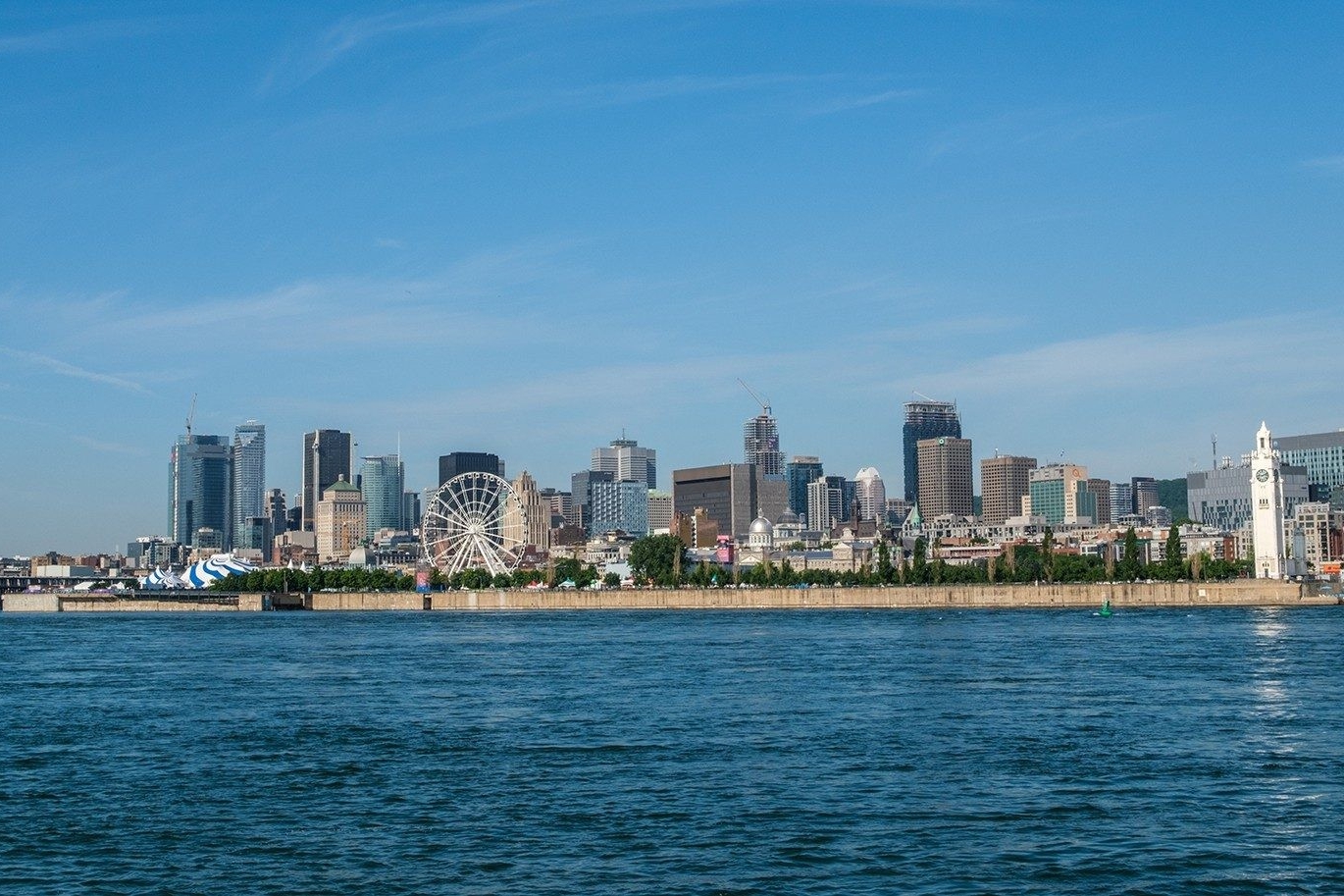A skyline of Montreal's Old Port as viewed from the river during the day shows buildings, and La Grande Roue de Montréal