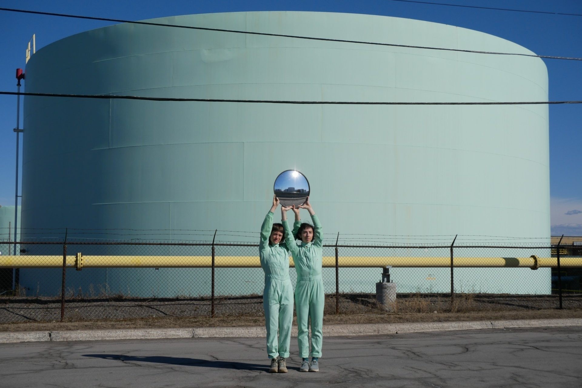 Two people where matching teal jumpsuits hold a circular mirror abovehead while standing in front of the Suncor refinery