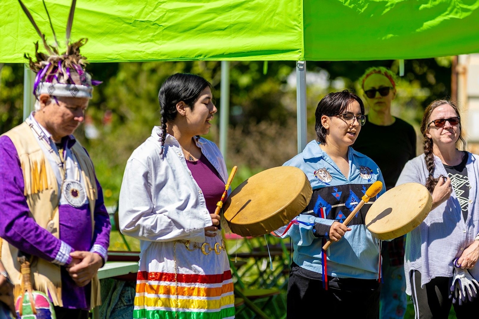 People standing outside on a bright sunny day, in traditional dress and celebrating  the opening of a community garden with drumming and song.