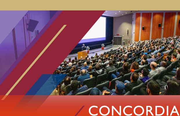 A large group of people seated in an auditorium, facing a presentation screen at the front, during a lecture or conference.