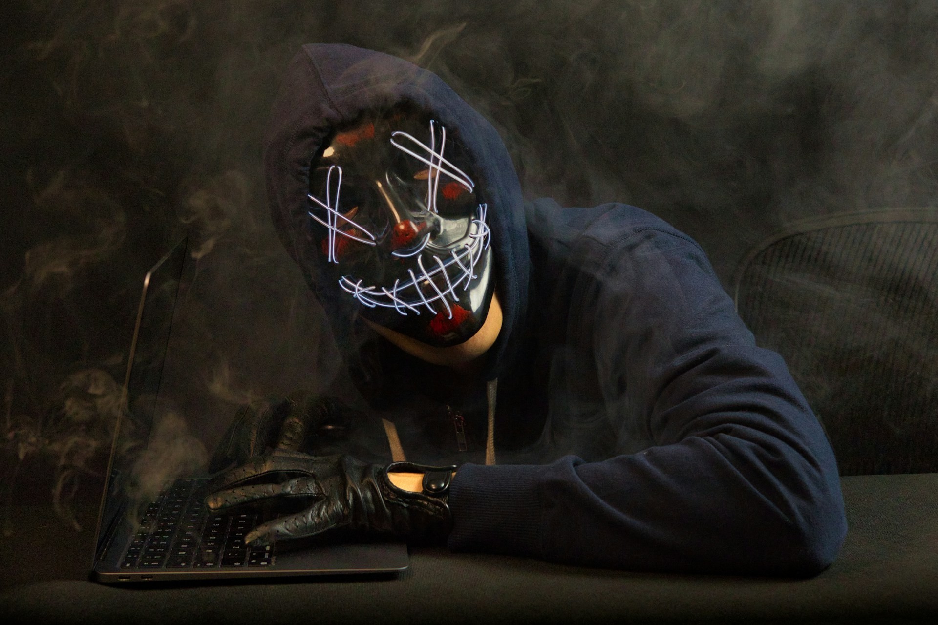 A figure wearing a black hoodie and mask with scribbles over the eyes and mouth faces the camera; a gloved hand rests hear a laptop.